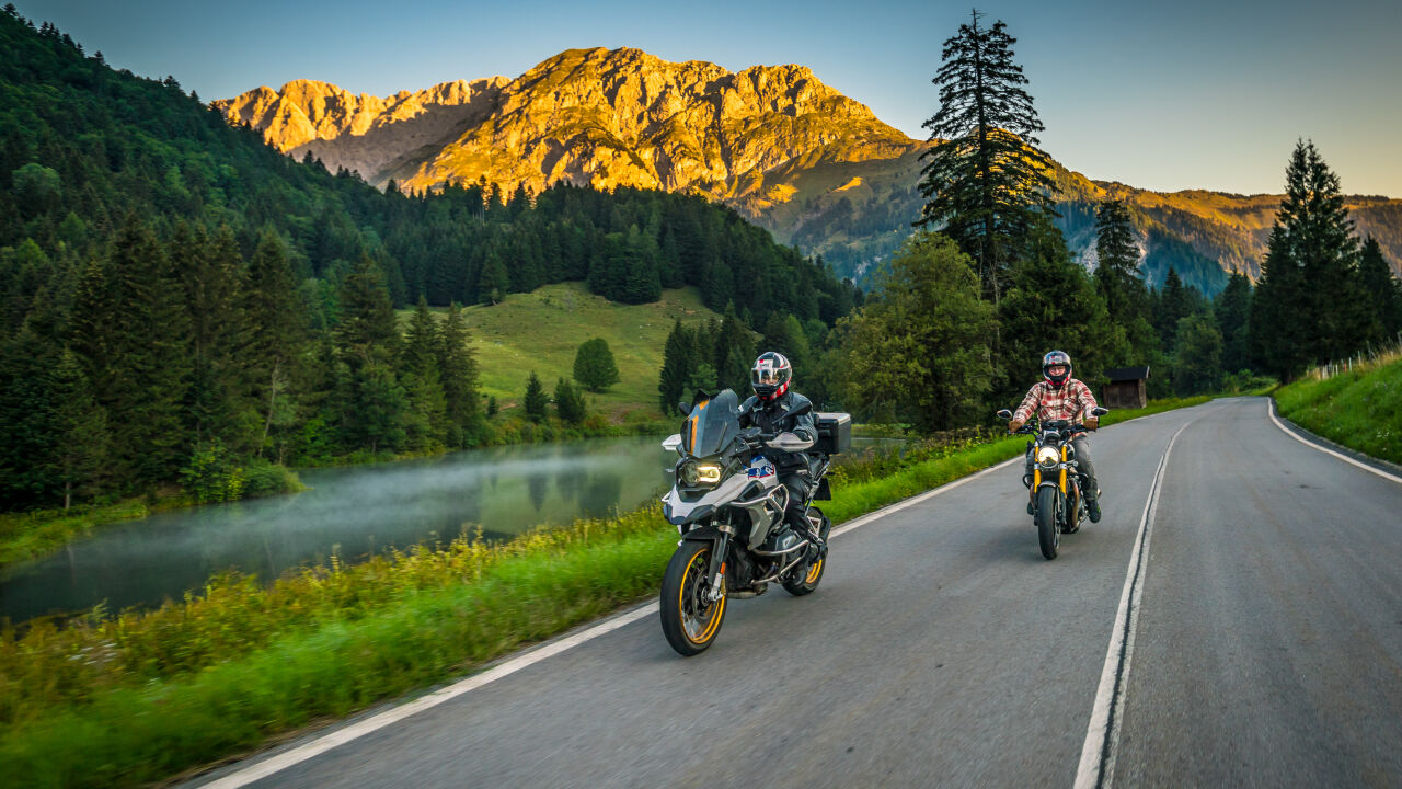 Zwei Motorräder auf einer Passstraße neben einem kleinen See, dahinter ein Felsmassiv im Morgenlicht
