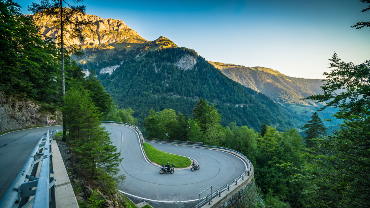 Zwei Motorräder in einer Kehre mit Leitplanke, dahinter ein bewaldetes Tal und Felsgipfel