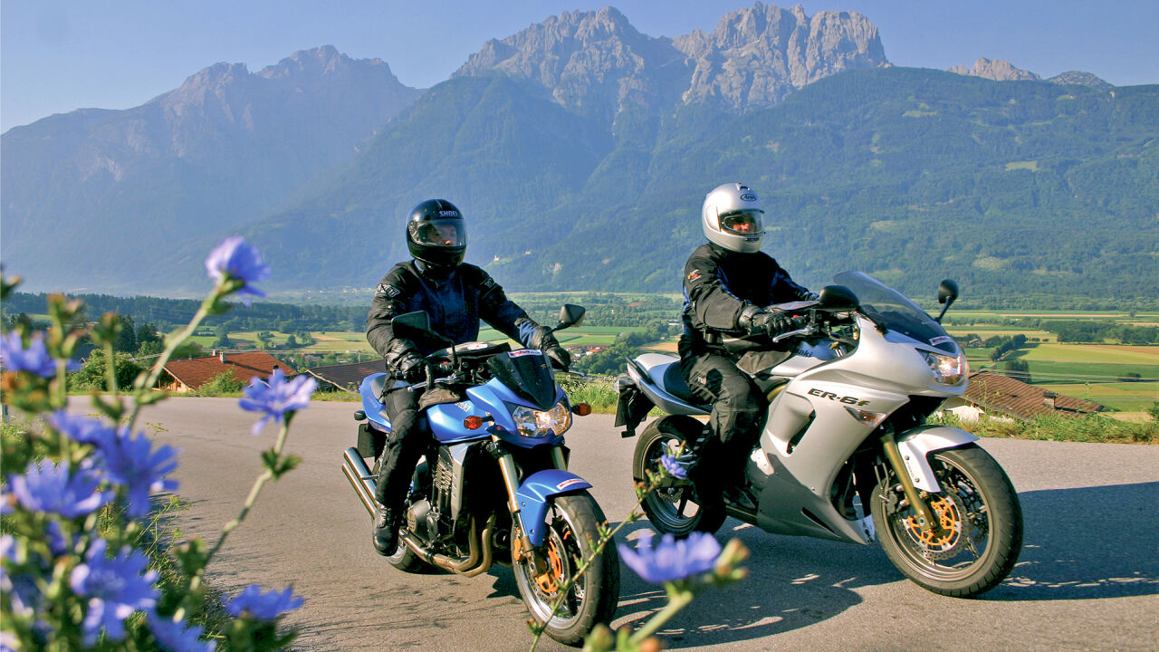 Zwei Motorräder an einer Passstraße mit Kornblumen, dahinter ein Talboden und ein Kalkmassiv